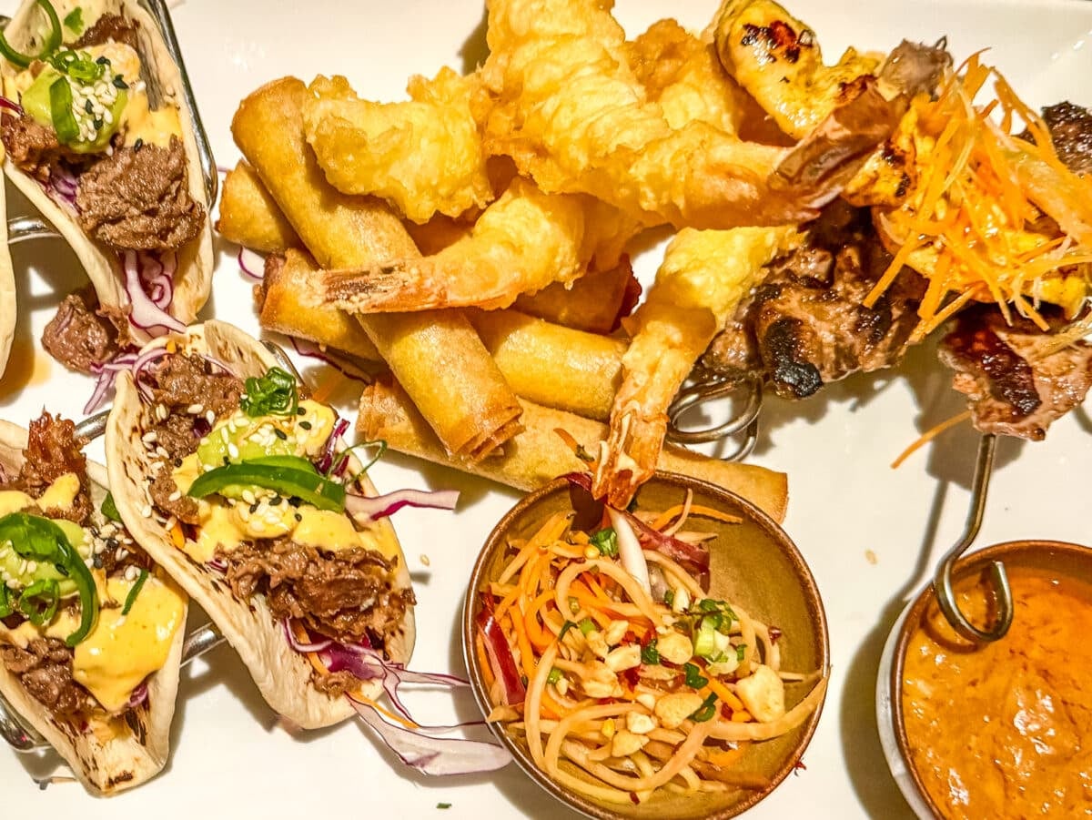 A plate with beef tacos, fried shrimp, spring rolls, grilled skewers, pickled vegetable salad, and a bowl of dipping sauce—perfect for enjoying on a Holland America cruise with teens.