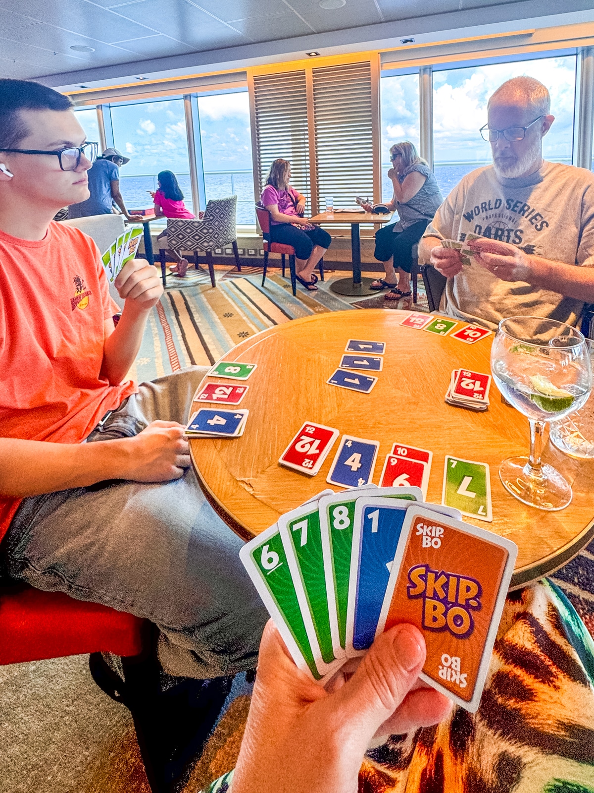 Three people sit at a round table near large windows on Holland America with teens, playing a game of Skip-Bo with colorful cards spread out on the table.