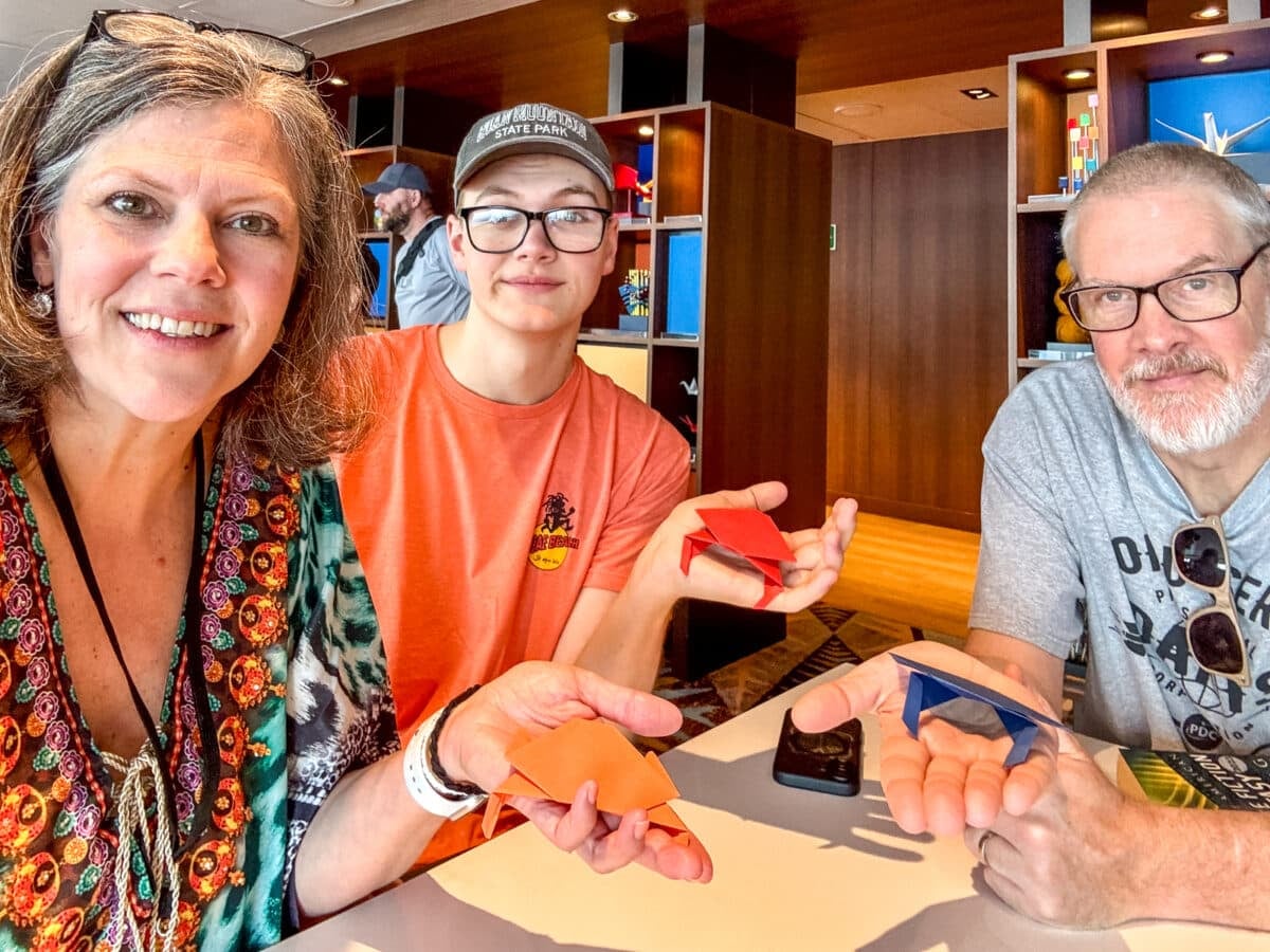 Three people sit at a table indoors, each holding a folded origami object, enjoying an activity perfect for Holland America with teens. Shelves with books and colorful objects are visible in the background.