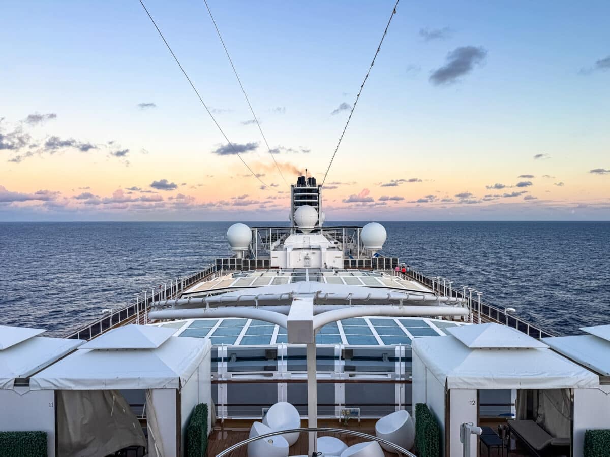 View from the upper deck of a Holland America cruise ship sailing on open water at sunset, with white cabins and seating visible in the foreground—a perfect setting for families cruising with teens.