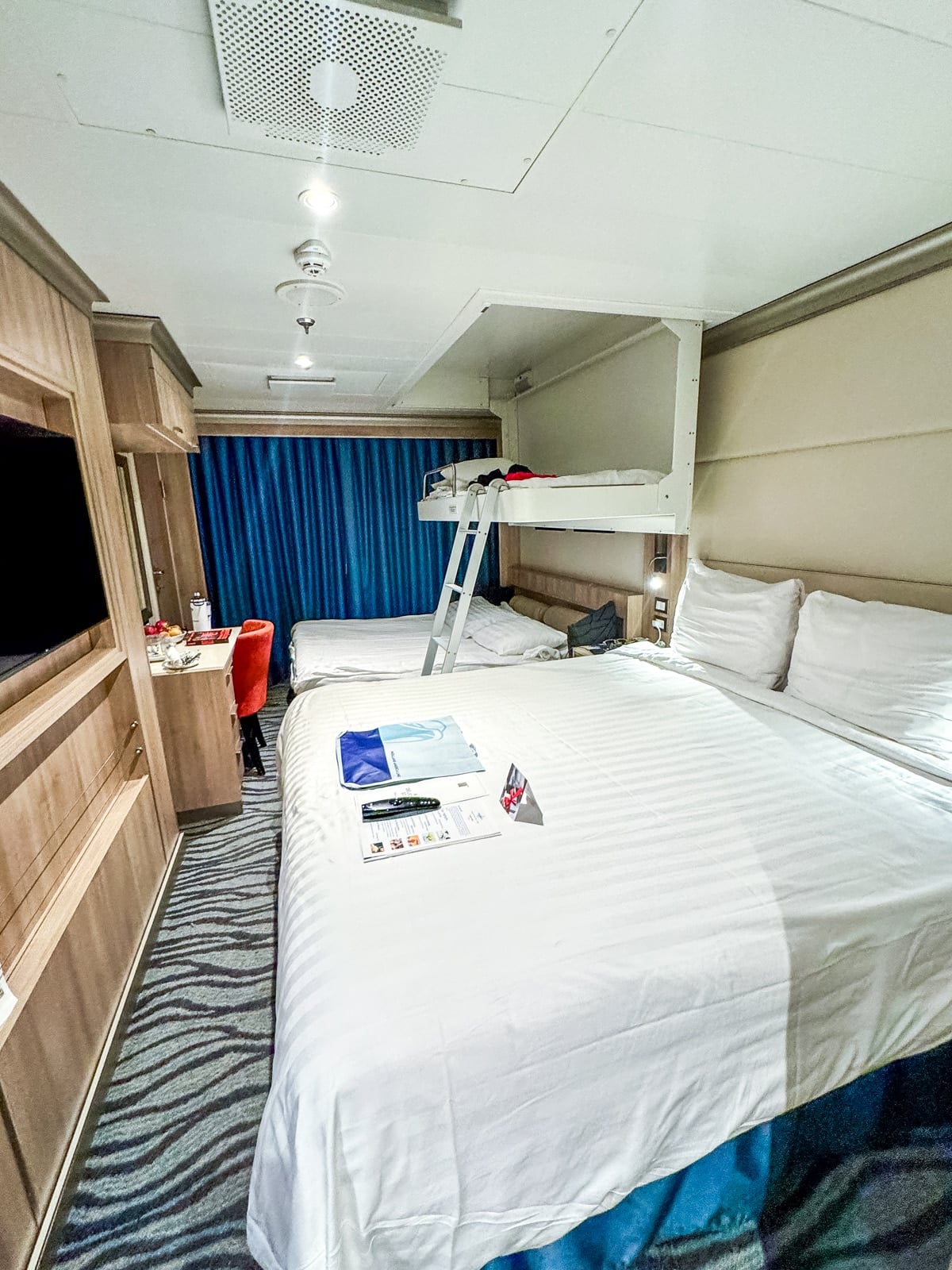 A compact cruise ship cabin on Holland America, perfect for families with teens, features a double bed, a bunk bed above, a desk with chair, TV, wardrobe, and blue curtains covering the window.