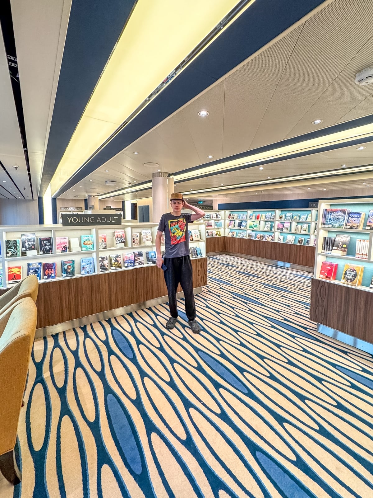 A person stands in a modern library's young adult section, surrounded by shelves filled with books on a patterned carpet, browsing titles popular with teens—much like those found on Holland America with teens aboard.
