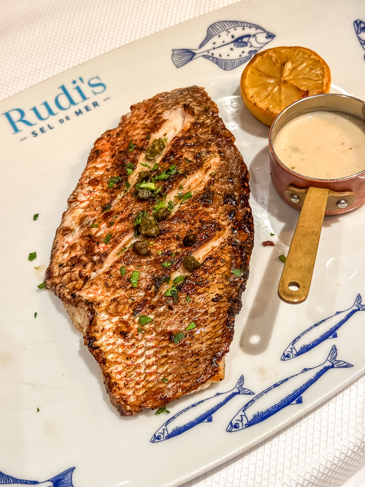 Grilled fish fillet garnished with herbs, served with a grilled lemon half and a small cup of sauce on a decorative plate labeled "Rudi’s Sel de Mer"—a delightful dish to enjoy while sailing Holland America with teens.