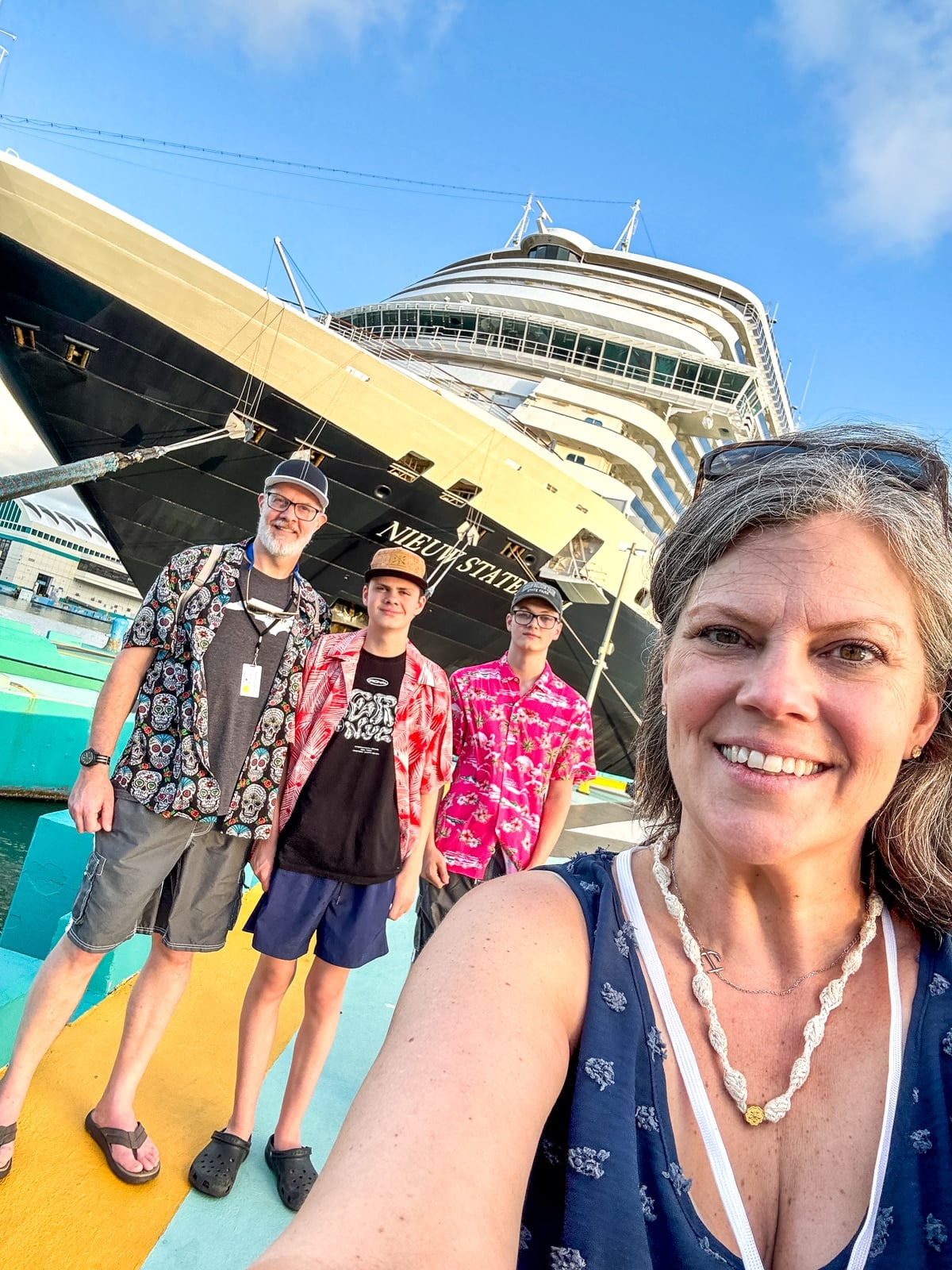 Four people pose for a selfie in front of a large Holland America cruise ship docked at a port, under a blue sky—an exciting adventure with teens awaits.