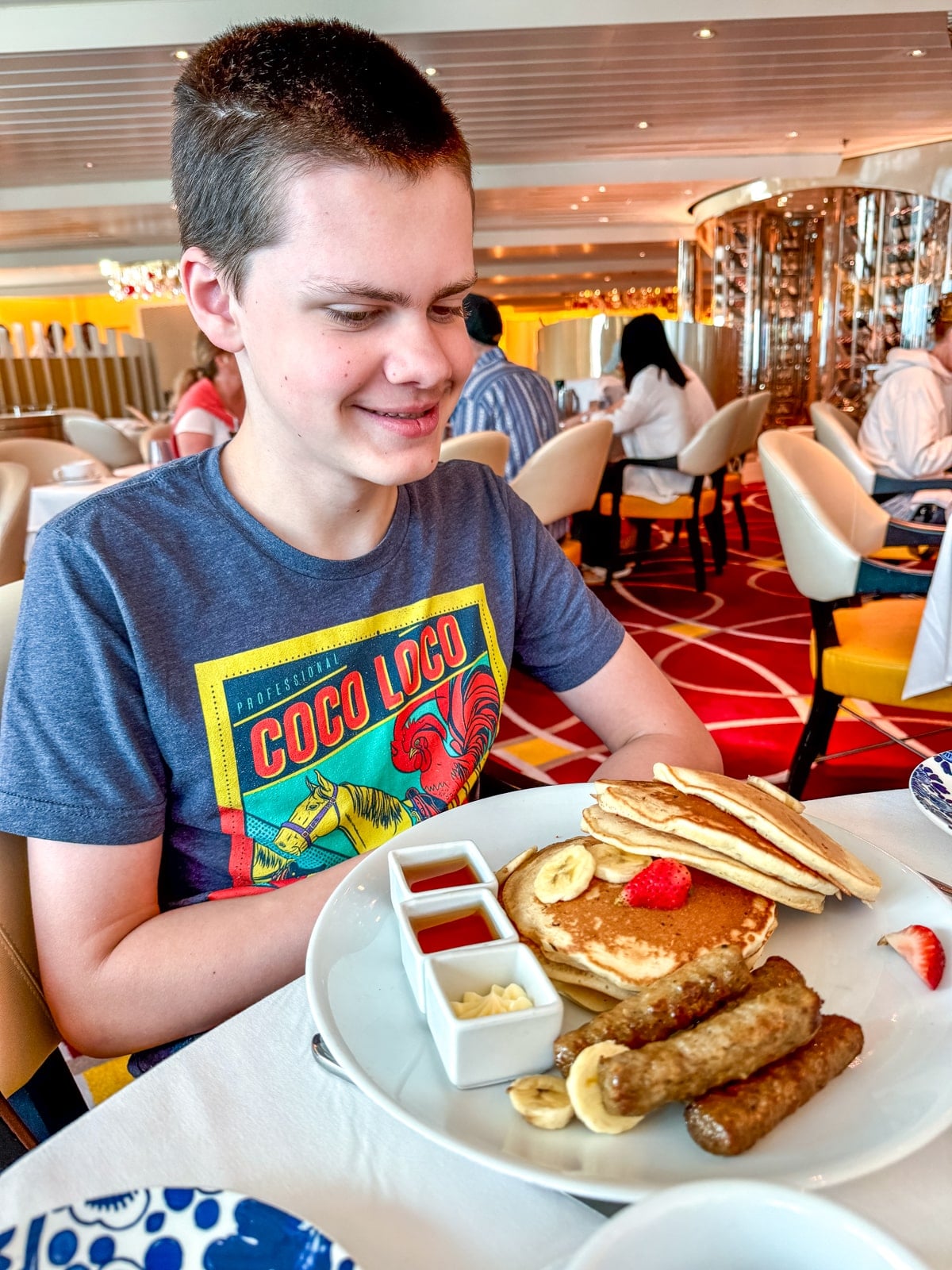 A boy with short brown hair smiles at a plate of pancakes, sausages, bananas, and syrup in a brightly lit restaurant aboard Holland America with teens.