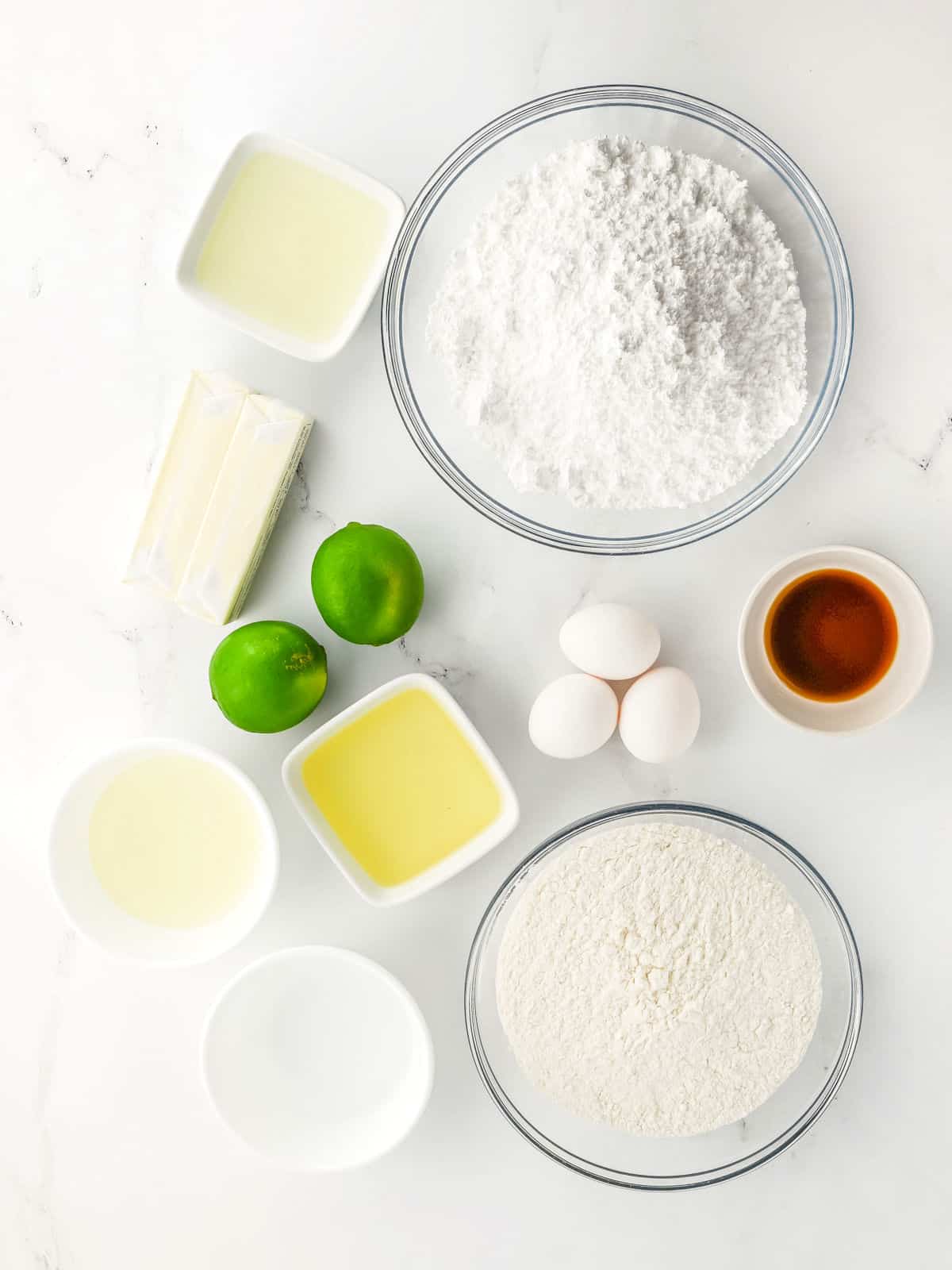 Various baking ingredients including flour, powdered sugar, eggs, limes, butter, oil, vanilla extract, and other liquids arranged in bowls on a white surface.