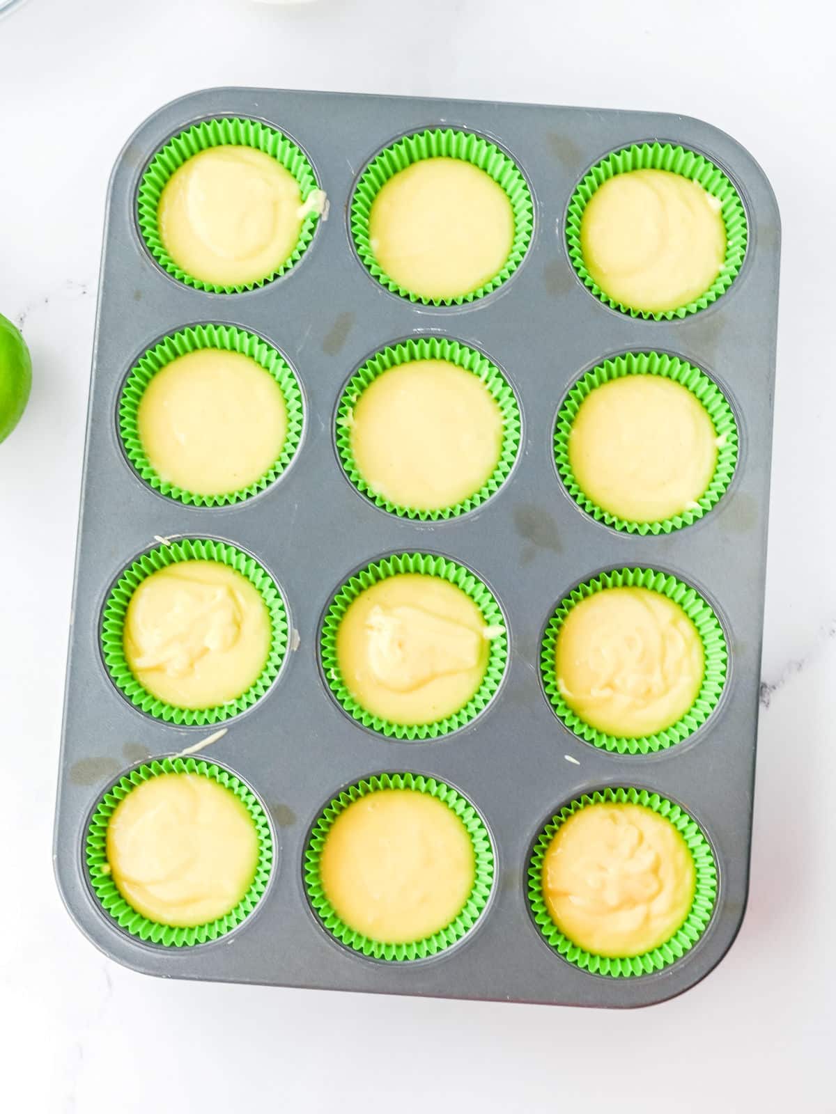 A muffin tin lined with green paper cups, each filled with yellow batter, sits on a white surface.