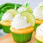 Vanilla cupcakes with white frosting, garnished with a lime slice and green-striped straw, in green paper liners on a wooden surface.