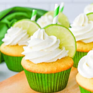 Vanilla cupcakes with white frosting, garnished with a lime slice and green-striped straw, in green paper liners on a wooden surface.