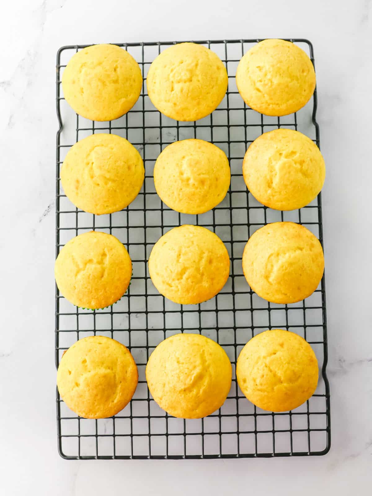 Twelve plain yellow muffins cooling on a black wire rack, placed on a white marble surface.