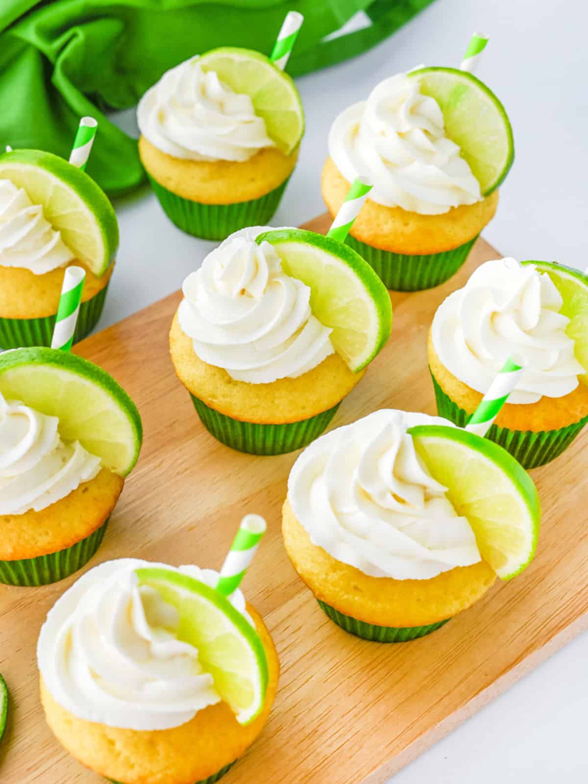 Cupcakes with white frosting, each topped with a lime wedge and a green-striped straw, arranged on a wooden board with a green cloth in the background.