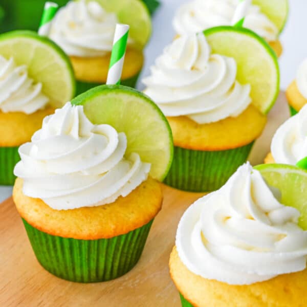 Several cupcakes with white frosting, each topped with a slice of lime and a green-striped paper straw, sit on a wooden surface.
