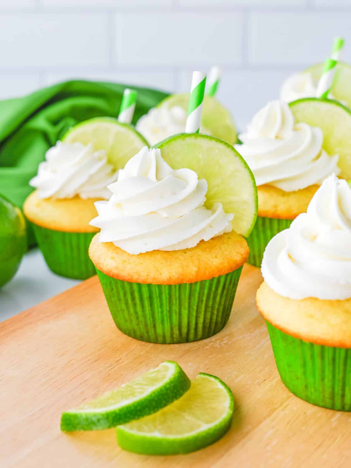 Cupcakes with white frosting, lime slices, and green-striped straws are arranged on a wooden surface, with extra lime slices in front and a green cloth in the background.