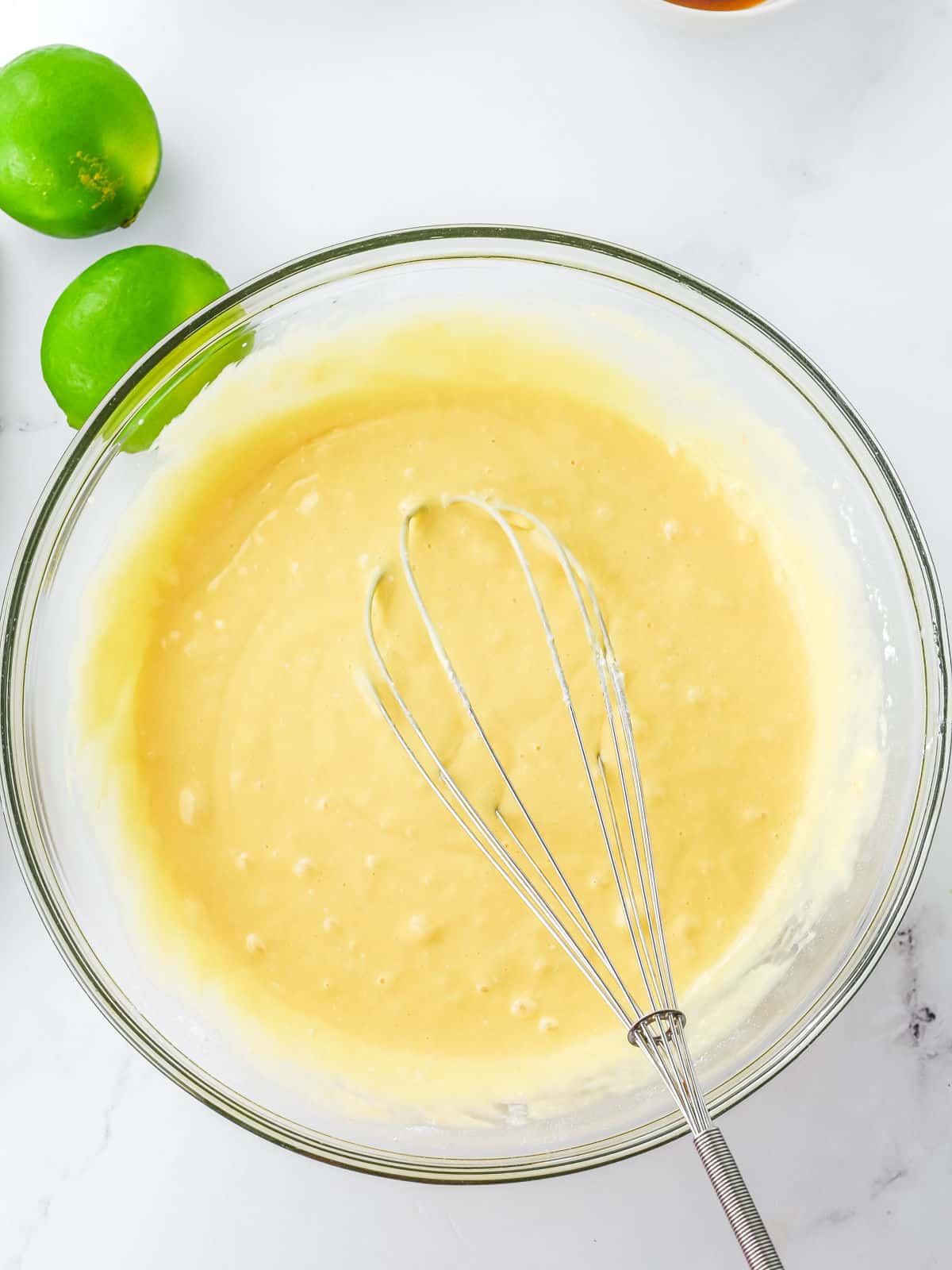A glass bowl of yellow cake batter with a metal whisk inside, placed on a white surface next to two whole limes.