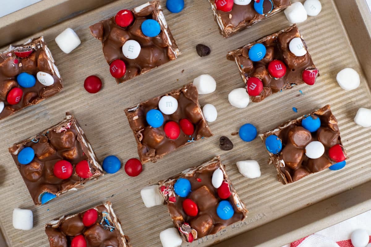 Nine chocolate dessert bars topped with red, white, and blue M&M candies are arranged on a baking sheet, surrounded by mini marshmallows and loose M&Ms.