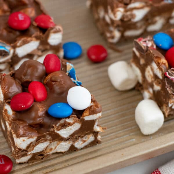 Squares of chocolate marshmallow treats topped with red, white, and blue candy-coated chocolates are arranged on a baking tray with loose candies and marshmallows nearby.