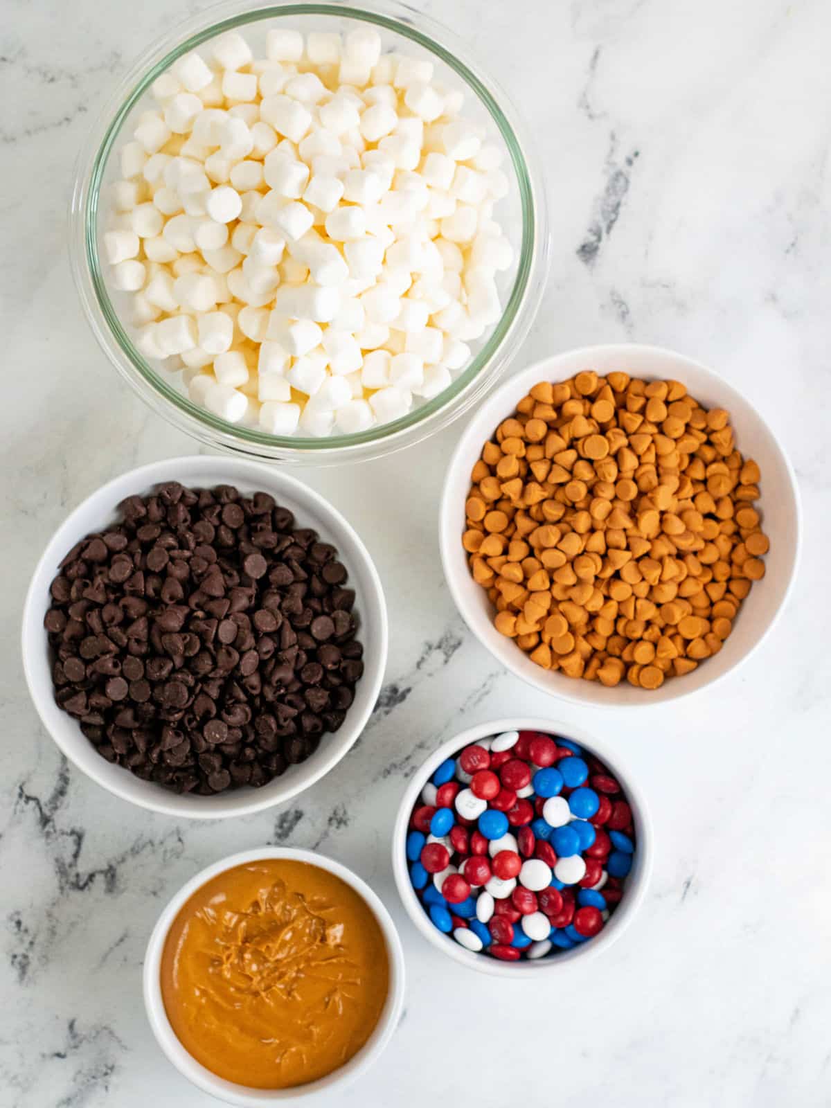 Five bowls on a marble surface containing mini marshmallows, butterscotch chips, chocolate chips, red-white-blue candies, and peanut butter.