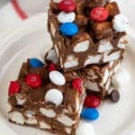 Three chocolate marshmallow bars topped with red, white, and blue candy-coated chocolates are stacked on a white plate, with a few extra candies and marshmallows scattered around.