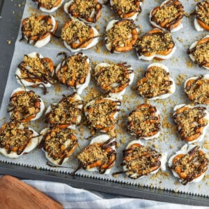 A baking tray lined with parchment paper holds clusters of pretzels topped with white coating, chocolate drizzle, and crumbly topping.