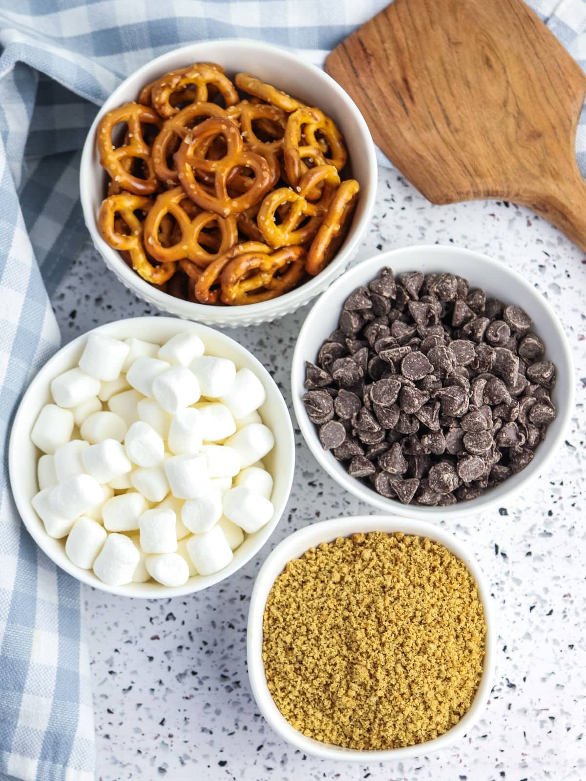 Four bowls containing pretzels, chocolate chips, mini marshmallows, and graham cracker crumbs are arranged on a speckled countertop next to a wooden spatula and a blue plaid cloth.