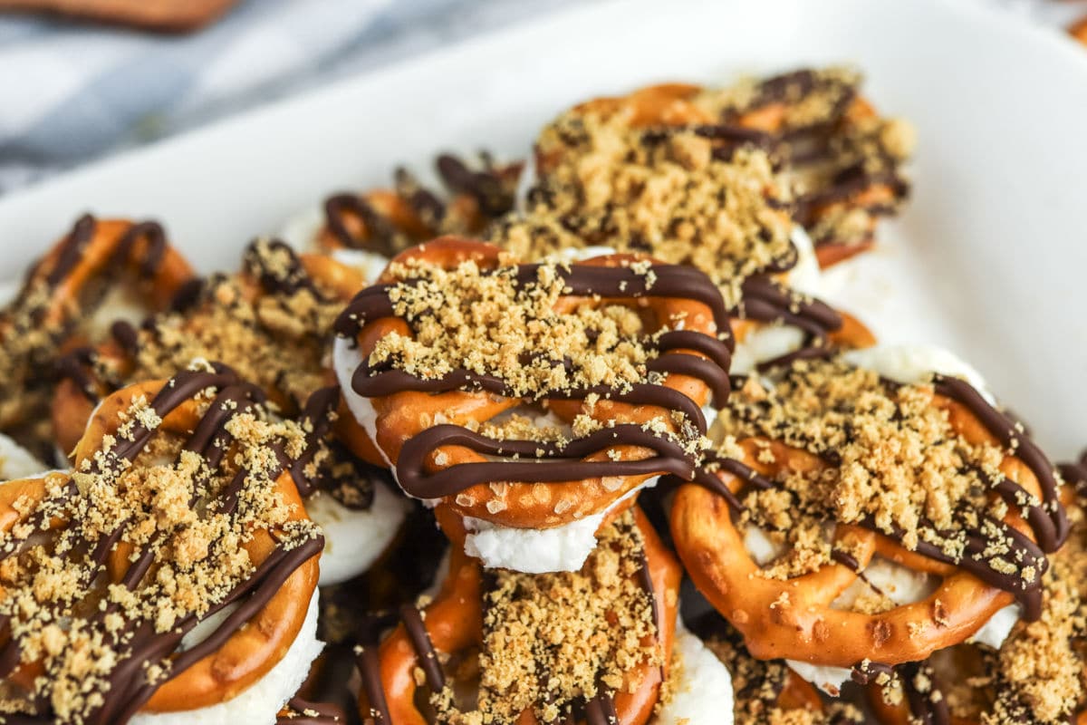 A close-up of pretzel sandwiches filled with marshmallow, topped with chocolate drizzle and crushed graham crackers on a white plate.