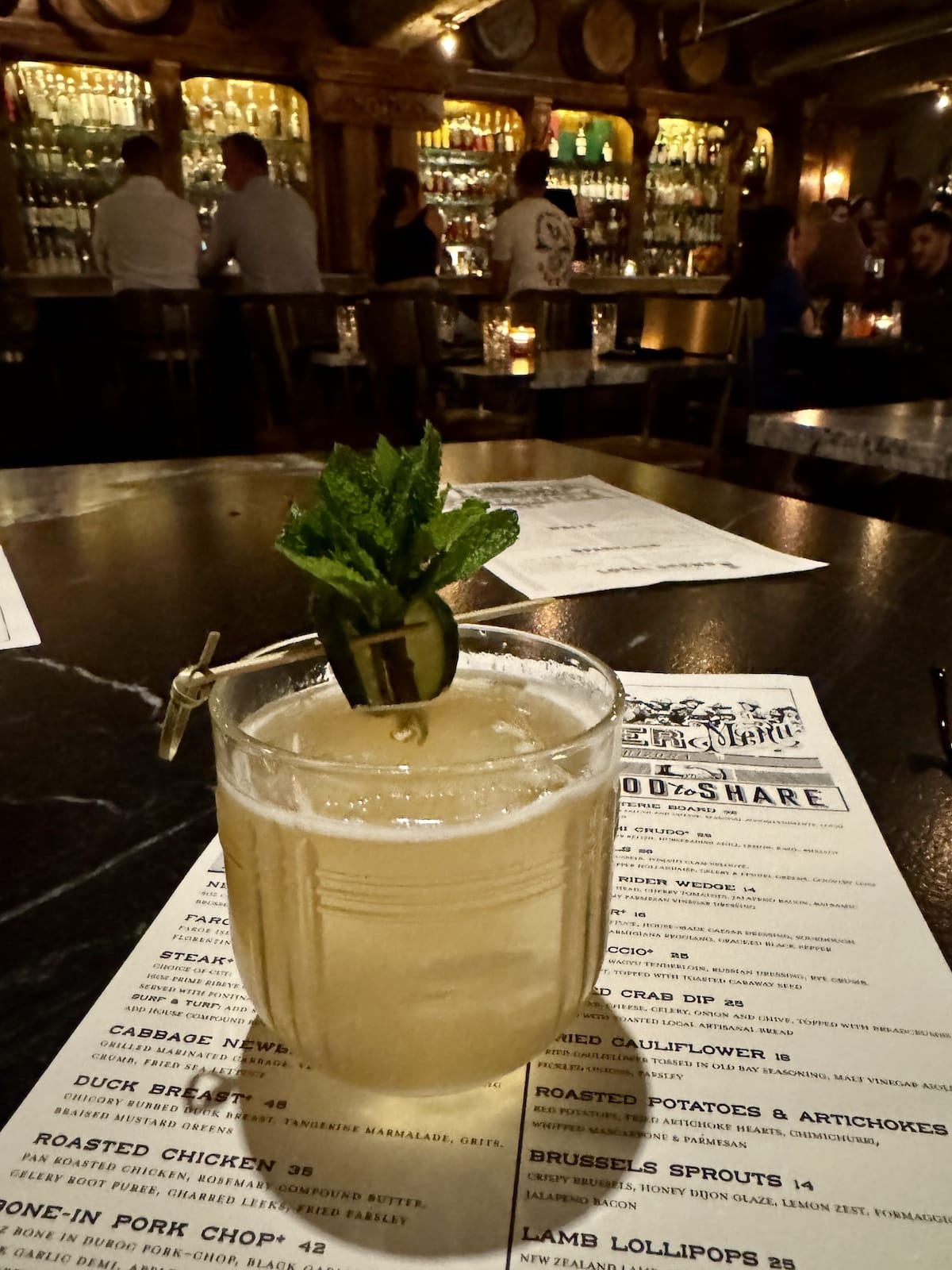A cocktail garnished with mint and cucumber sits on a restaurant menu on a dimly lit bar table, with patrons and shelves of liquor bottles in the background.