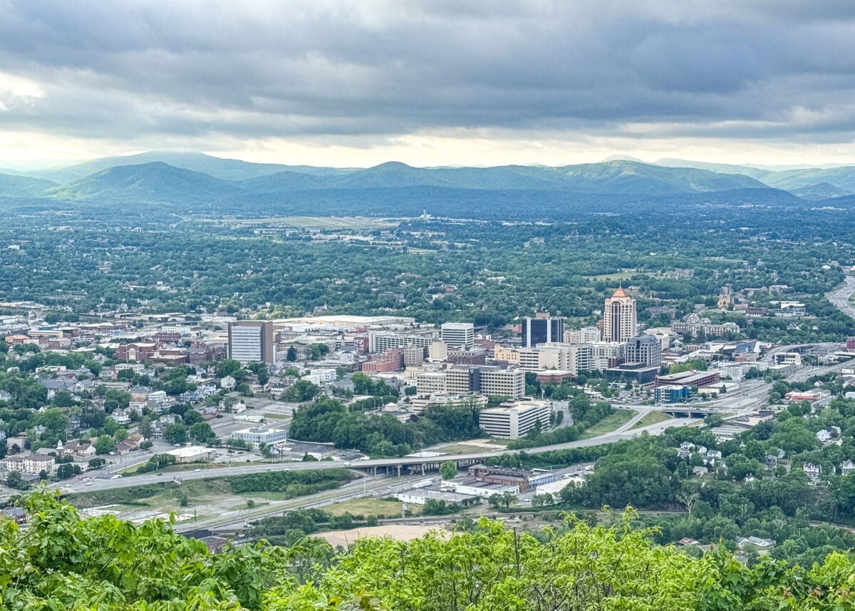 Aerial view of a city with clustered buildings, highways, and surrounding greenery&mdash;discover Things to Do in Roanoke VA, set against distant blue mountains under a cloudy sky.