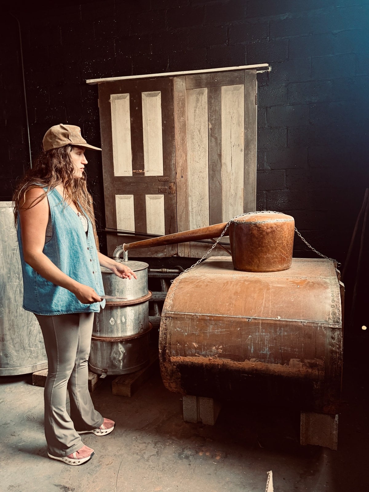 A woman wearing a cap stands beside an old, rustic still in a dimly lit room with wooden barrels and a weathered door&mdash;capturing the charm of local distilleries among the unique things to do in Roanoke VA.