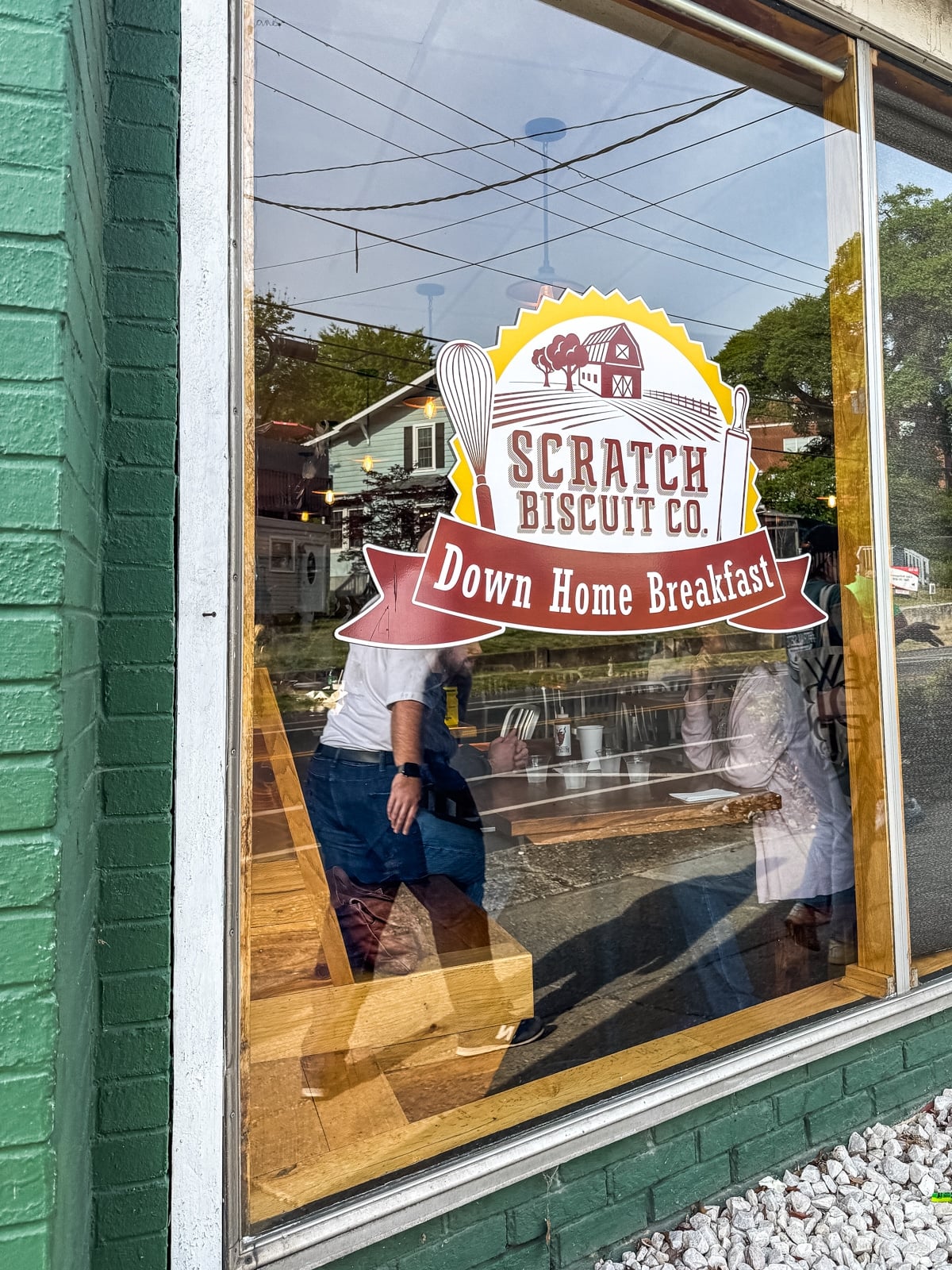 Large window displays the "Scratch Biscuit Co. Down Home Breakfast" logo; people are seated inside at wooden tables. Reflections show surrounding buildings and power lines&mdash;a cozy spot among the top Things to Do in Roanoke VA.