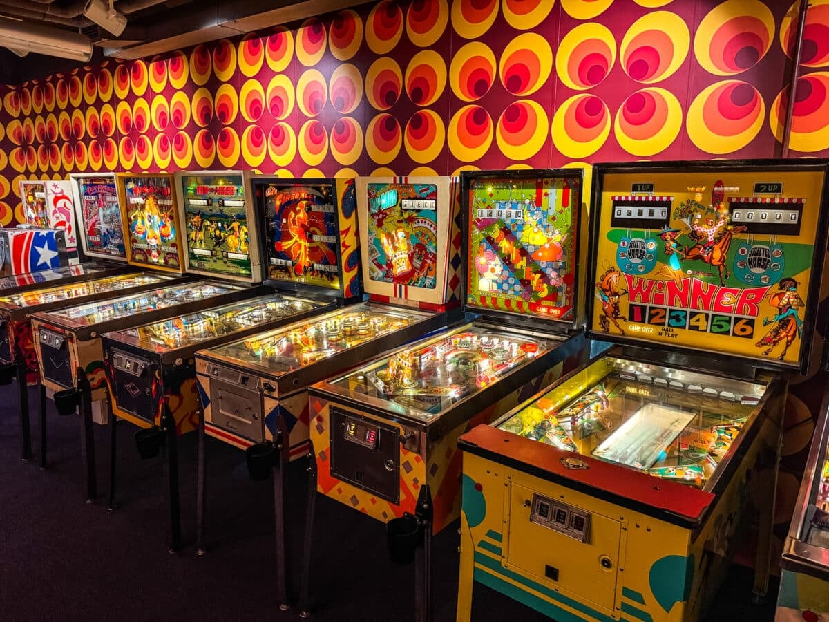 A row of vintage pinball machines in an arcade, set against brightly colored retro-patterned wallpaper&mdash;one of the nostalgic things to do in Roanoke VA for arcade and pinball enthusiasts.