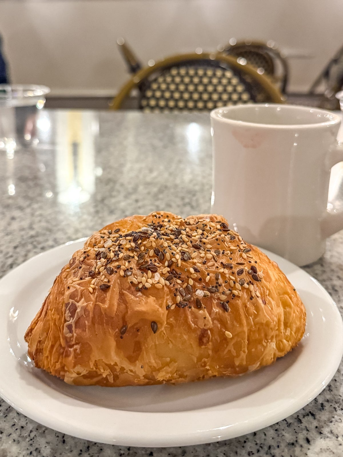 A seeded croissant on a white plate sits beside a white ceramic mug atop a gray marble table&mdash;a cozy cafe moment and one of the delightful things to do in Roanoke VA.