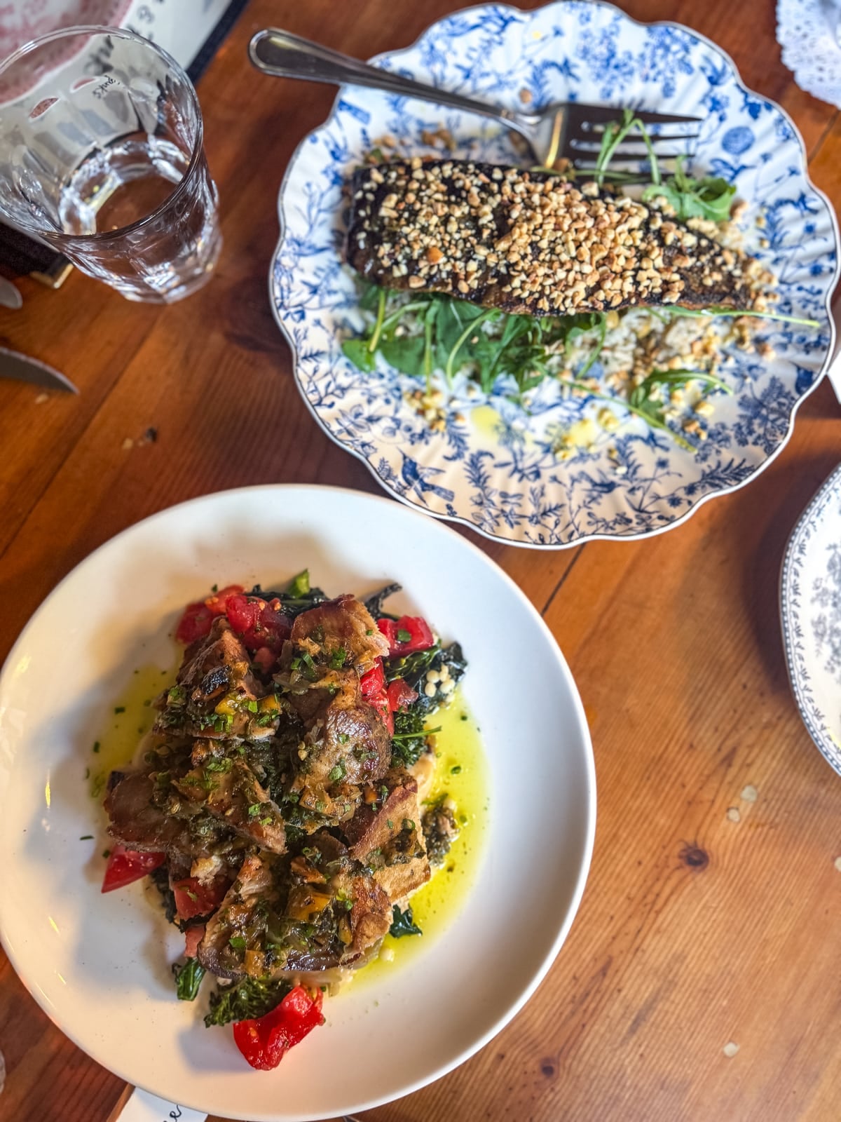 Two plates of food on a wooden table: one with grilled meat and vegetables in sauce, the other with a nut-crusted fish fillet on greens, plus a glass of water&mdash;a delicious highlight among Things to Do in Roanoke VA.