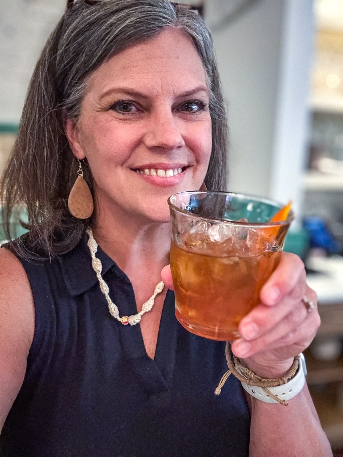 A woman with gray hair and a sleeveless black top smiles while holding up a glass of iced beverage with an orange garnish, enjoying one of the refreshing things to do in Roanoke VA.