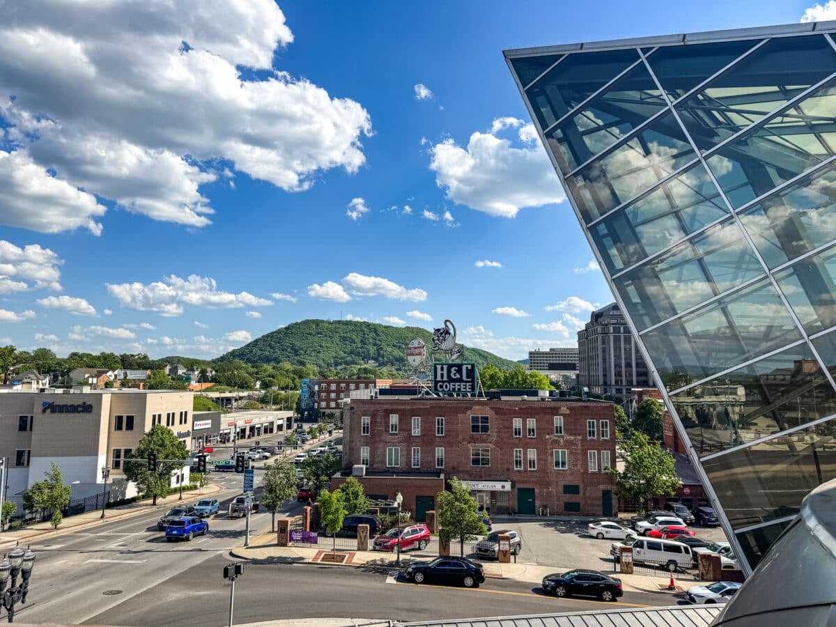 Street view of a city with a glass building on the right, a red brick coffee shop in the center&mdash;perfect for those exploring Things to Do in Roanoke VA&mdash;cars on the road, and a green hill under a blue sky with scattered clouds.