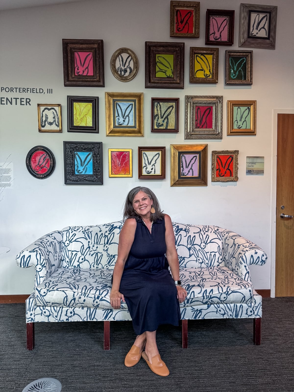 A woman sits on a white couch with a blue bunny pattern, in front of a wall displaying many colorful bunny prints&mdash;capturing the quirky charm you&rsquo;ll find among the unique things to do in Roanoke VA.