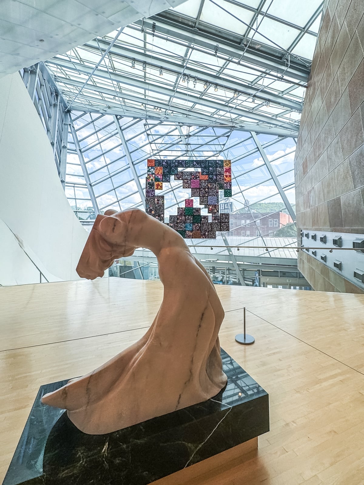 A marble sculpture of a headless, draped figure is displayed on a black pedestal in a modern gallery with large glass windows&mdash;one of the unique things to do in Roanoke VA, complemented by a colorful quilt artwork in the background.