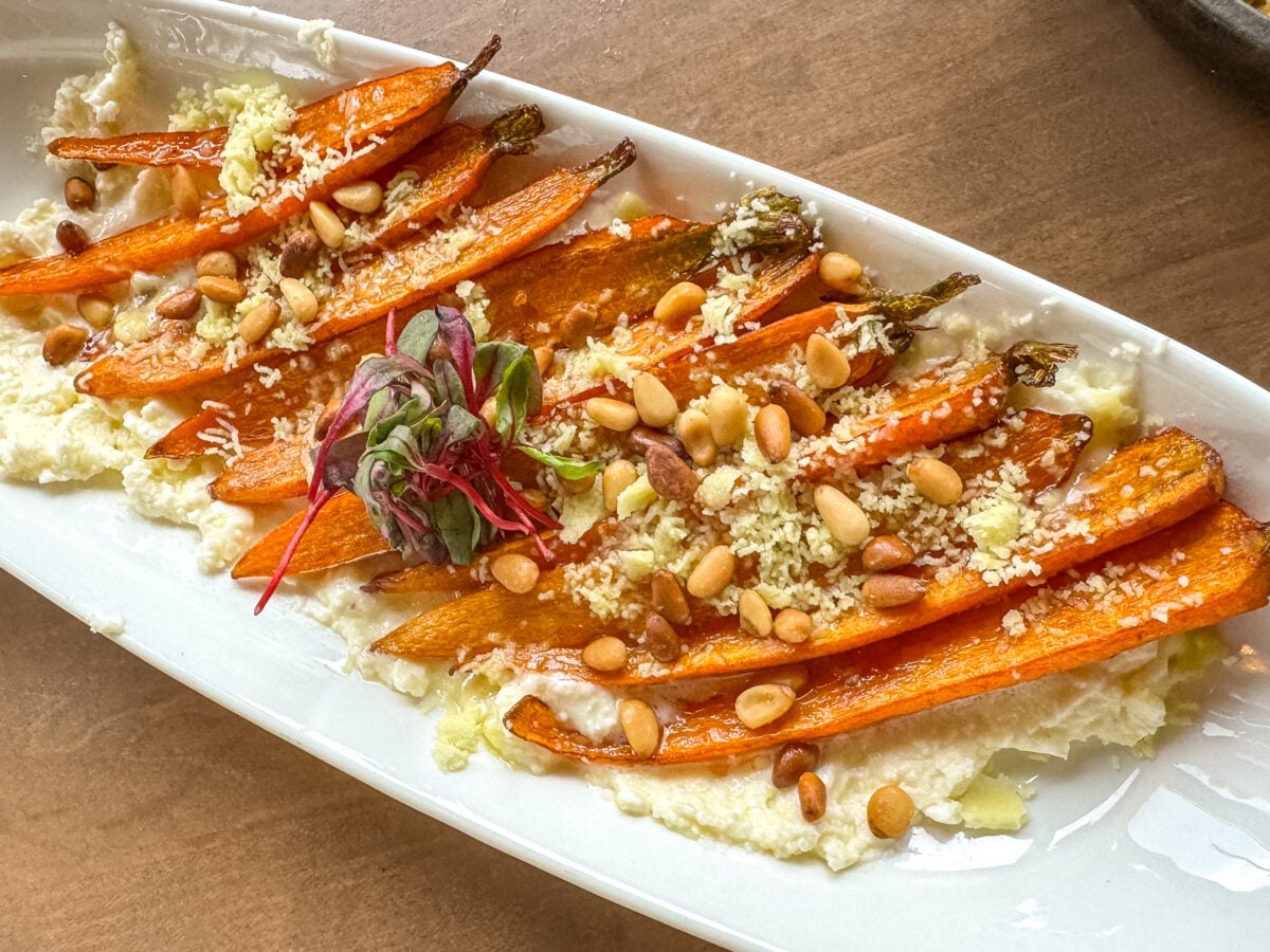 Roasted carrots on a bed of creamy cheese, topped with pine nuts, grated cheese, and microgreens&mdash;an unforgettable dish to enjoy while exploring Things to Do in Roanoke VA, served elegantly on a white oval plate.