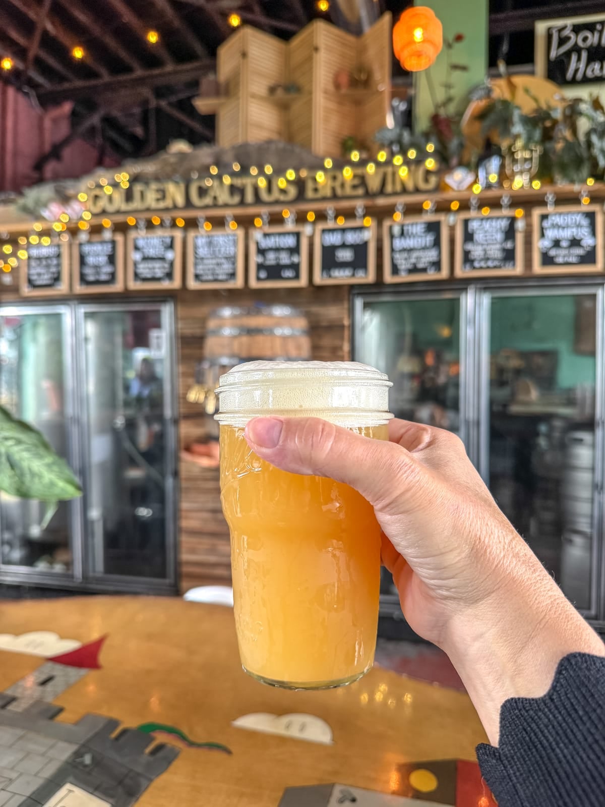 A hand holds a pint of hazy yellow beer in front of the bar at Golden Cactus Brewing, one of the top things to do in Roanoke VA, with menu boards and decor visible in the background.