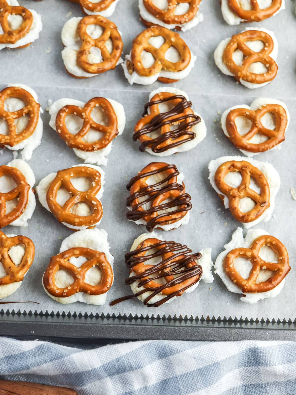 Rows of pretzel pieces on a baking tray with a white filling, some drizzled with chocolate, placed on parchment paper.