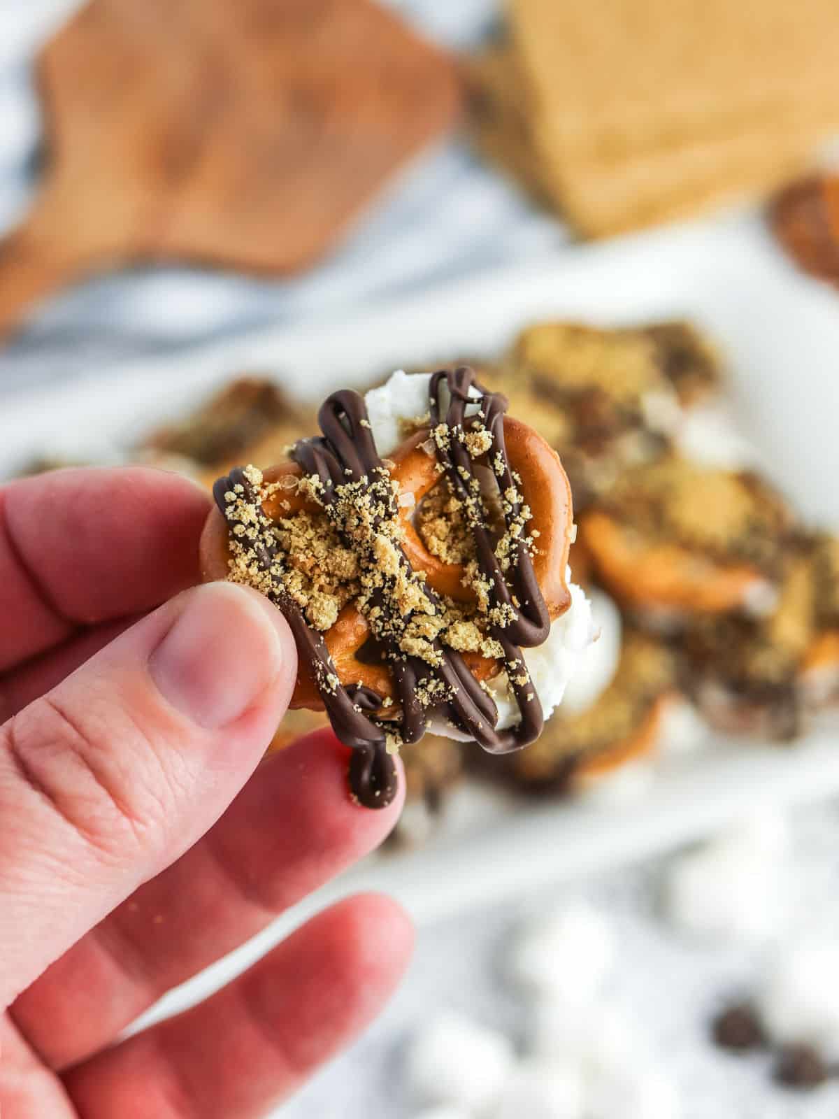 A hand holding a pretzel topped with melted marshmallow, chocolate drizzle, and crushed graham crackers, with more treats blurred in the background.