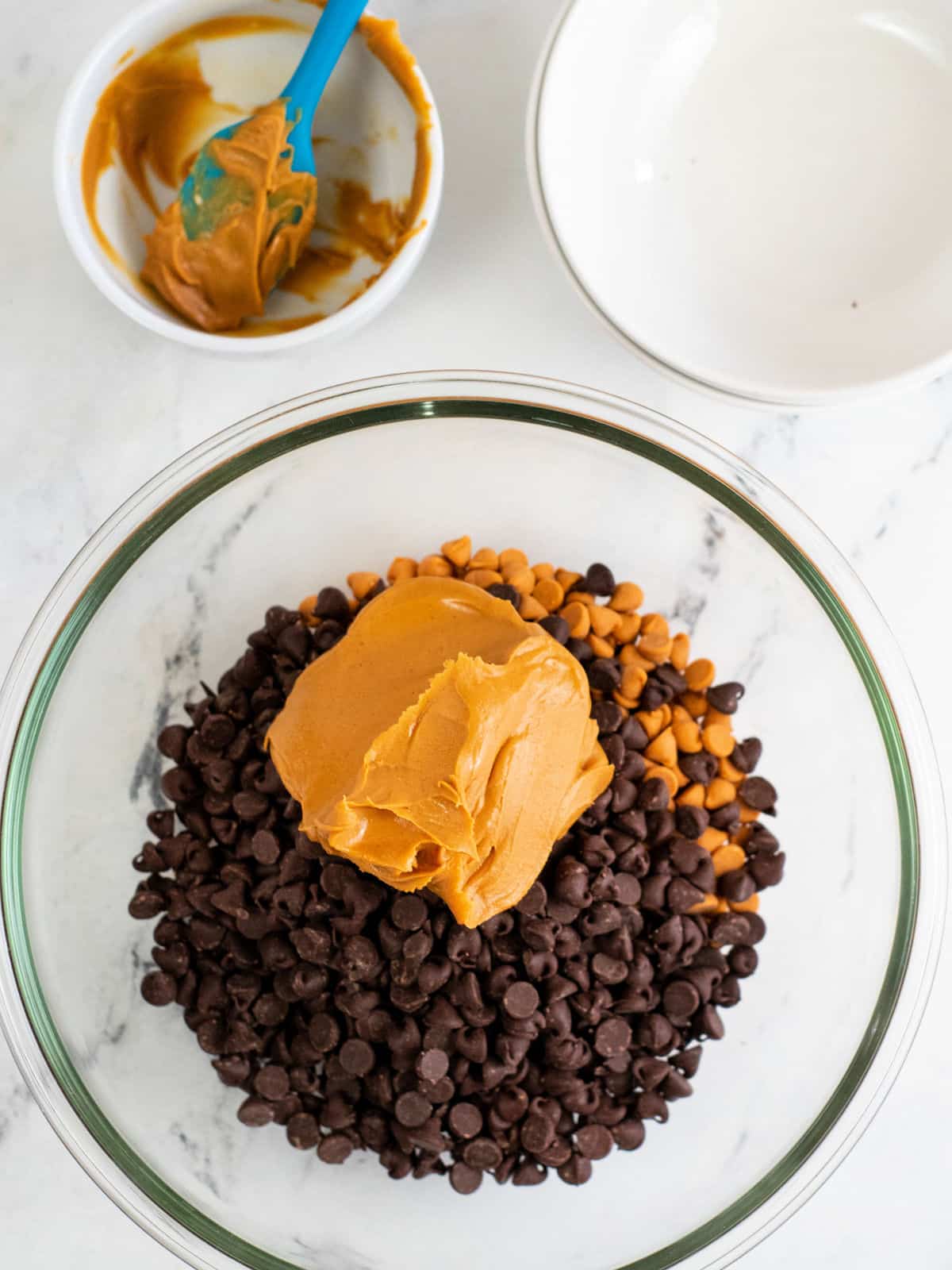 A glass bowl filled with chocolate chips, peanut butter chips, and a large dollop of peanut butter. Nearby are two small empty bowls, one with a blue spatula and peanut butter residue.