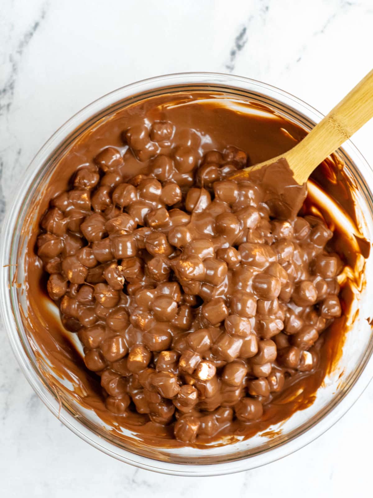 A glass bowl filled with melted chocolate and mini marshmallows being mixed with a wooden spoon on a marble surface.