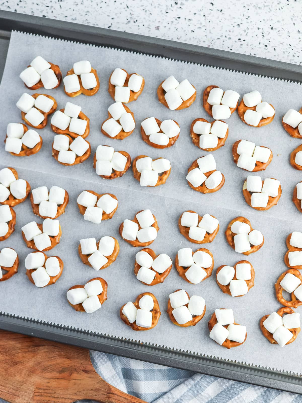 A baking sheet lined with parchment paper holds pretzel rounds topped with melted chocolate and several mini marshmallows on each.
