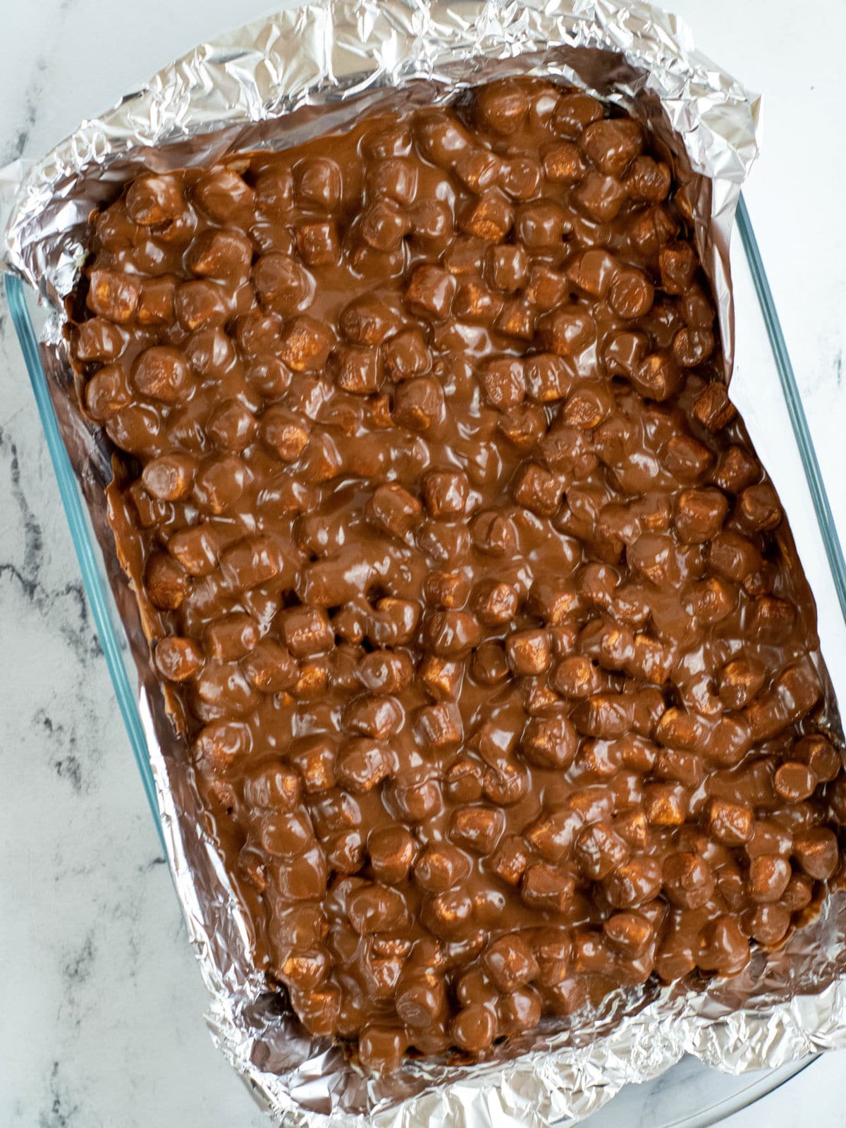 A glass baking dish lined with foil, filled with melted chocolate and mini marshmallows mixed together.