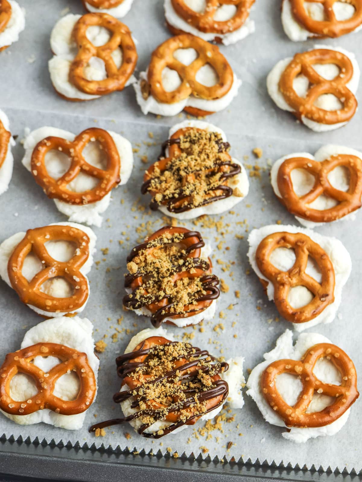 Pretzels topped with a white filling are arranged on parchment paper, with three in the center drizzled with chocolate and sprinkled with crushed toppings.