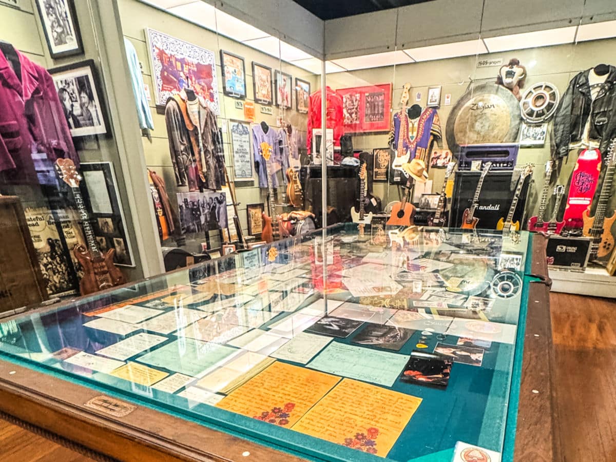 A glass display case with letters and photos sits in front of a wall filled with musical instruments, clothing, and memorabilia in a museum exhibit.