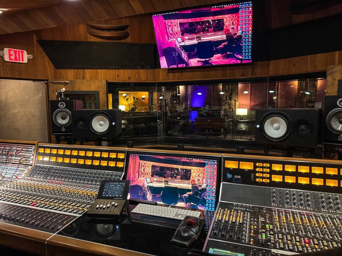 Professional recording studio with a large audio mixing console, studio monitors, computer screens, and interior view of a soundproof room through a glass window.