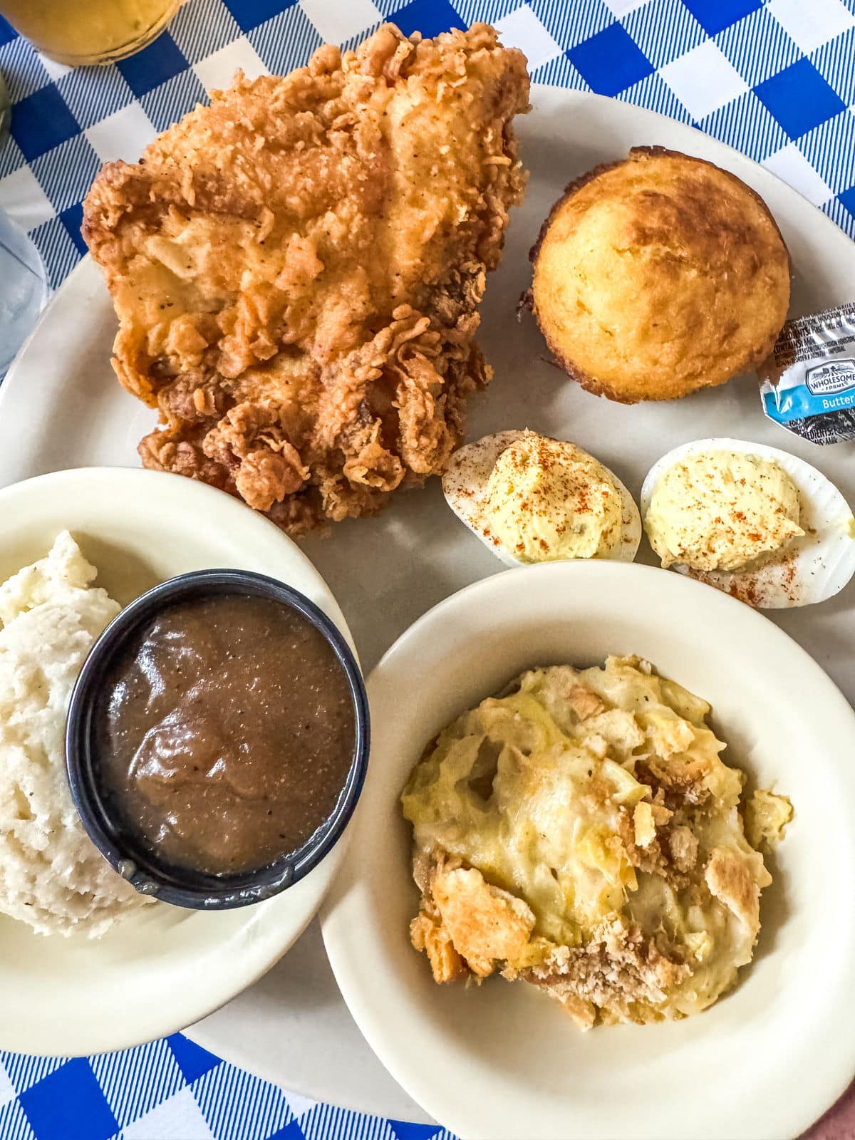 Plate with fried chicken, cornbread, deviled eggs, mashed potatoes with gravy, and a serving of casserole on a blue and white checkered tablecloth.