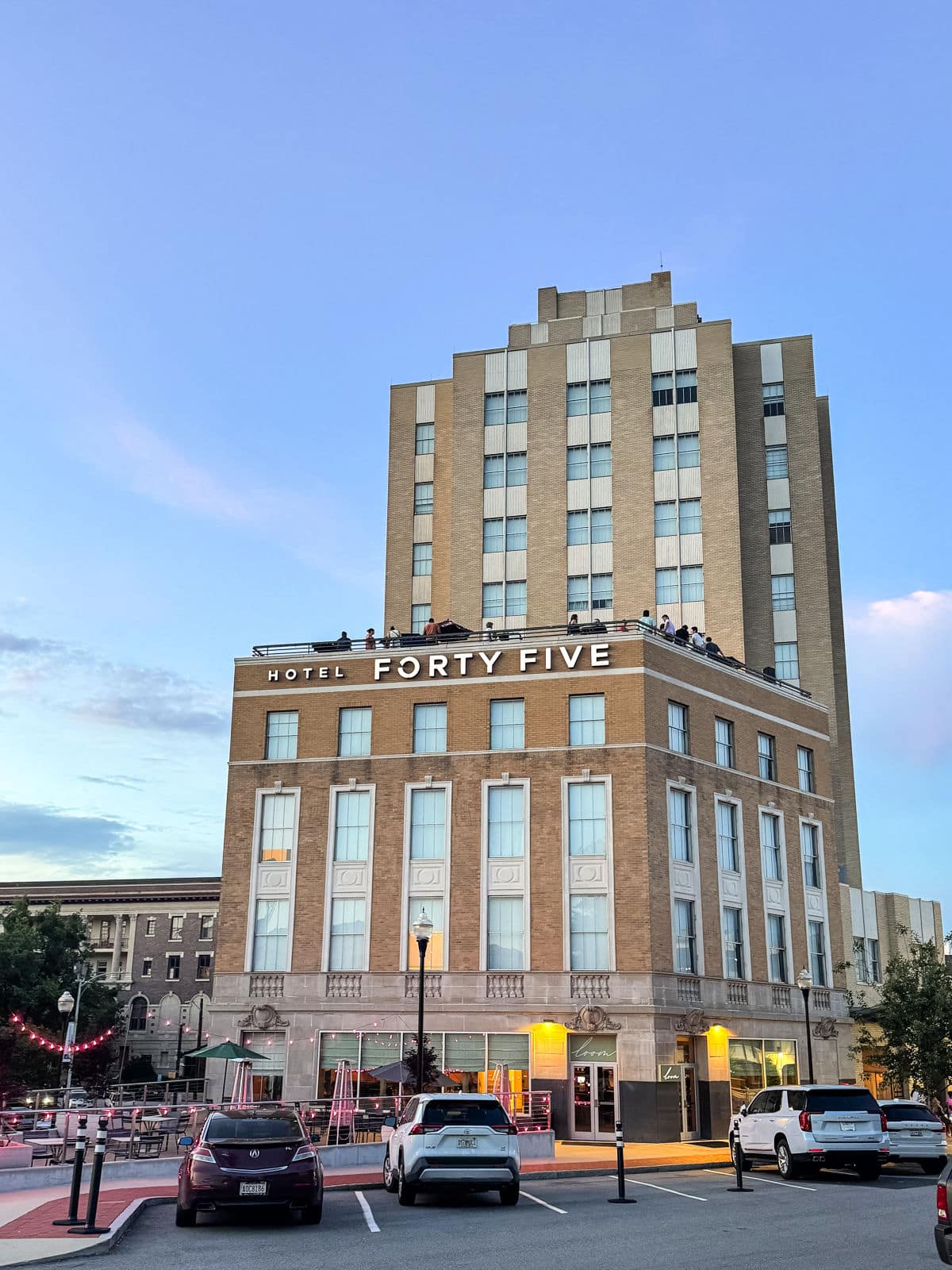 A multistory tan brick building labeled &ldquo;Hotel Forty Five&rdquo; with cars parked in front and people on a rooftop terrace at sunset.