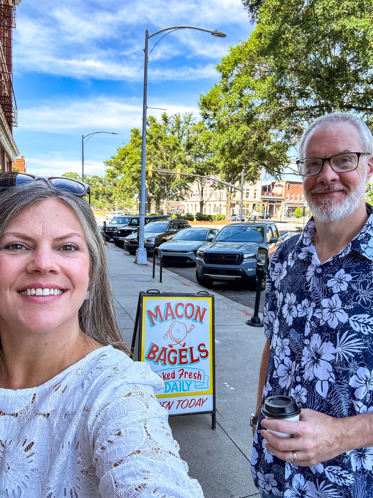Two people stand on a sidewalk in front of a &ldquo;Macon Bagels&rdquo; sign on a sunny day, with parked cars and trees in the background.